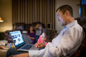 Sebastian Africano studies on his laptop in his living room with his daughter peeking at the screen