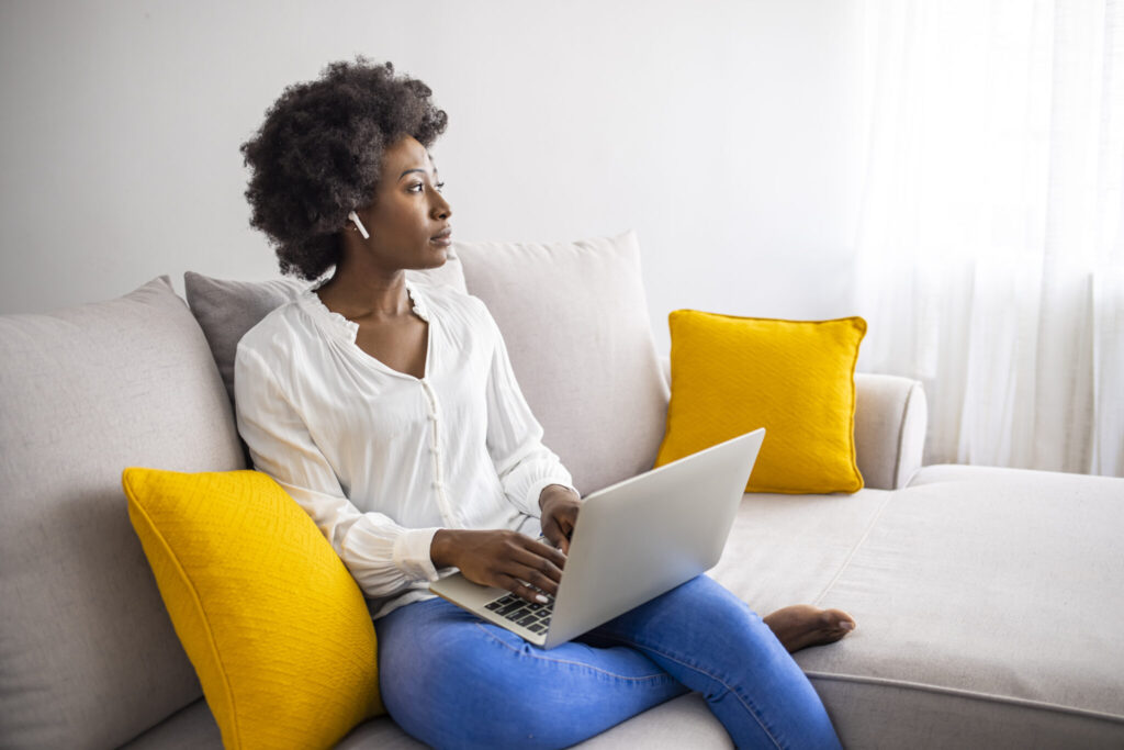 Young woman lying on her living room sofa at home wearing wireless earbuds and working on a laptop. Smiling young woman sitting in her living room at home wearing wireless earbuds and working