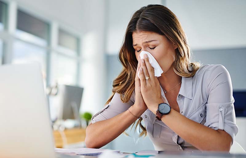 A woman sitting at a desk in the office blows her nose. The image illustrates the link between pay and health and wellness.